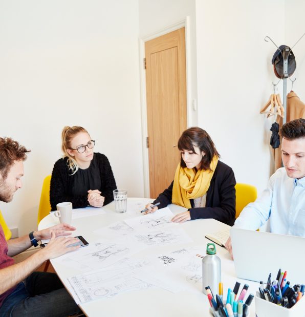 Four people at a business meeting around a table spread with papers, one man using a tablet and one on a laptop.